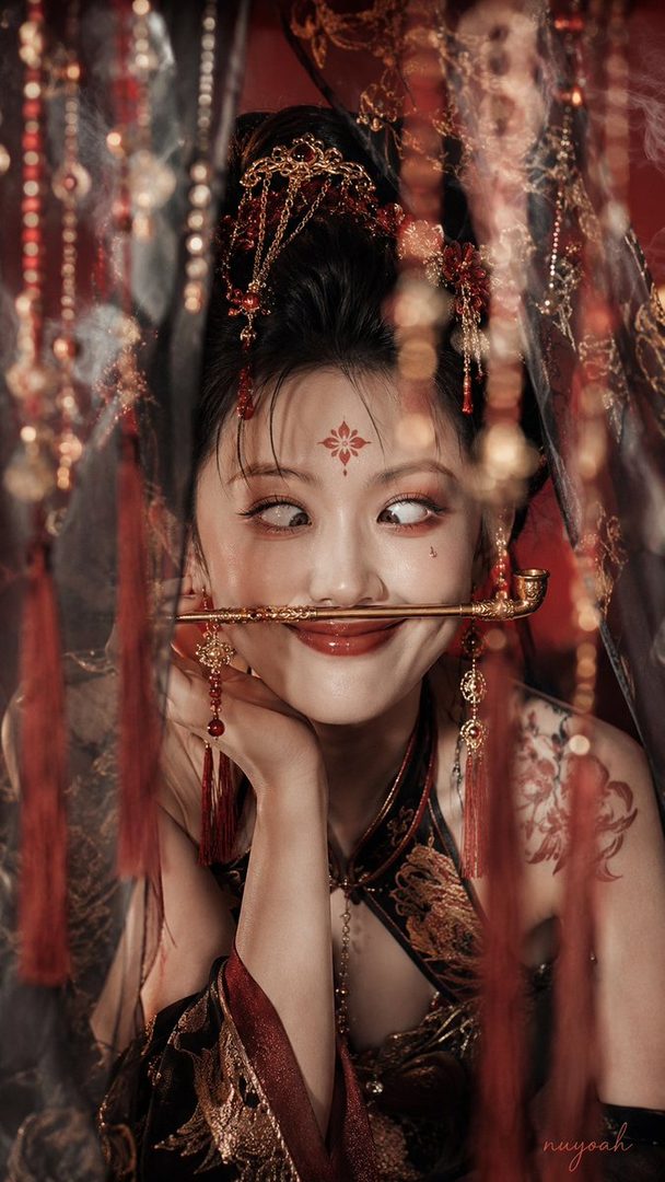 A woman in traditional Chinese attire with elaborate hair ornaments and makeup, holding a golden pipe in her mouth, with a playful and slightly mischievous expression.
