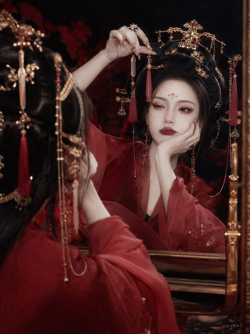 A woman in traditional red Chinese attire with elaborate hair ornaments looks into a mirror, posing elegantly.