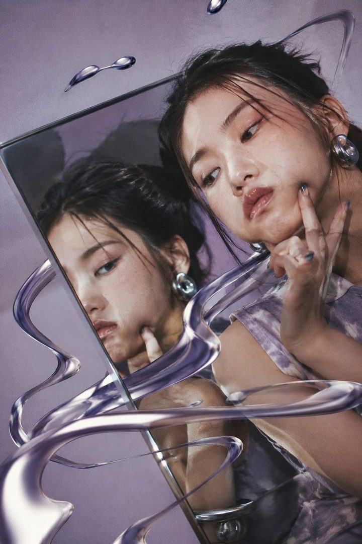 A young East Asian woman with dark hair pulled back poses in front of a mirror, her reflection creating a distorted double portrait. Abstract purple liquid metal swirls frame the image.