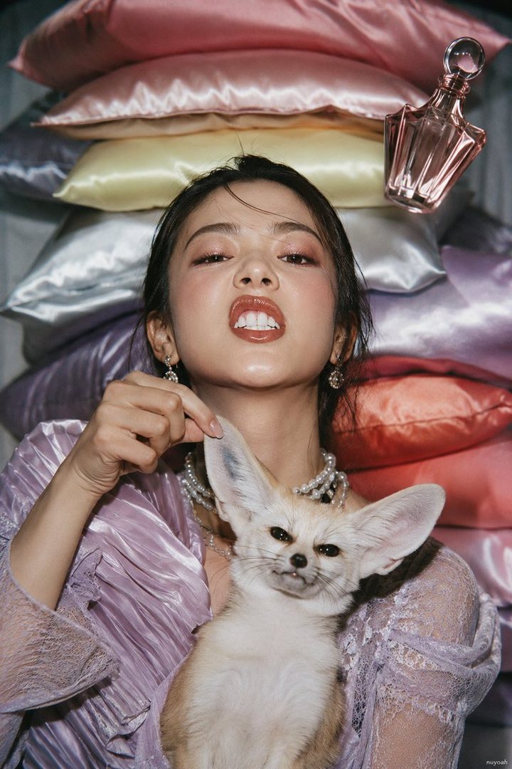 A young East Asian woman with dark hair, wearing a pearl necklace and a lilac blouse, playfully holds a fennec fox while posing in front of stacked satin pillows with a perfume bottle.