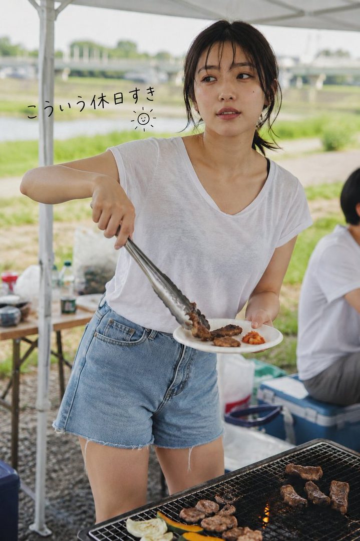 A young woman with dark hair tied in a ponytail, wearing a white t-shirt and denim shorts, is grilling meat at an outdoor barbecue by a river.
