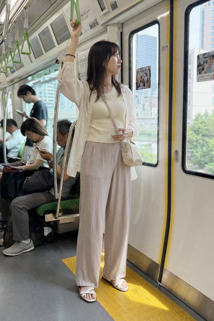 A young woman with dark brown hair stands in a subway train, holding onto a strap and looking out the window, dressed in a cream top, white cardigan, and beige wide-leg pants.