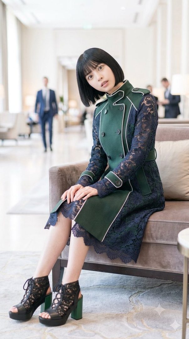 A young woman with short black hair and bangs, wearing a dark green and navy lace dress, sits on a sofa in a hotel lobby.