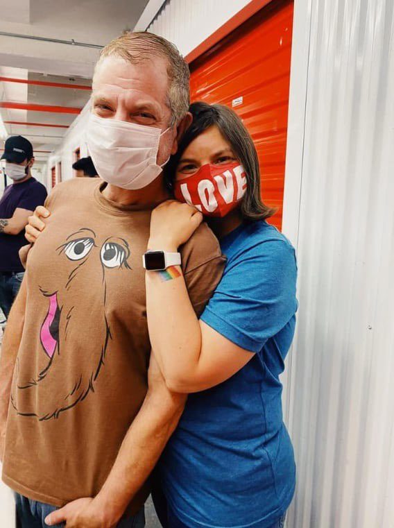 A couple wearing face masks, embracing each other. The woman has a red mask with 'LOVE' written on it, and the man wears a brown t-shirt with a cartoon character.
