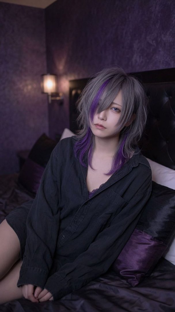 A young woman with purple-streaked grey hair, wearing a dark unbuttoned shirt over a lace top, poses suggestively on a bed in a dimly lit room.