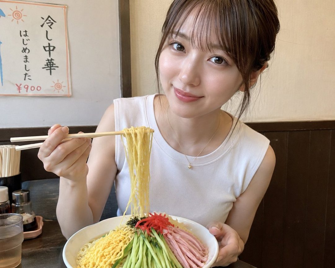 A young East Asian woman with brown hair and bangs, wearing a white sleeveless top, smiles as she holds chopsticks with noodles, about to eat a bowl of cold ramen.