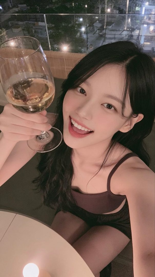 A young woman with long black hair smiles brightly while holding a glass of white wine, wearing a dark camisole, at an outdoor evening gathering.