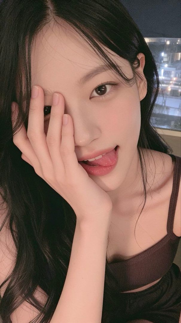 A young woman with long black hair playfully sticks out her tongue while covering part of her face with her hand.
