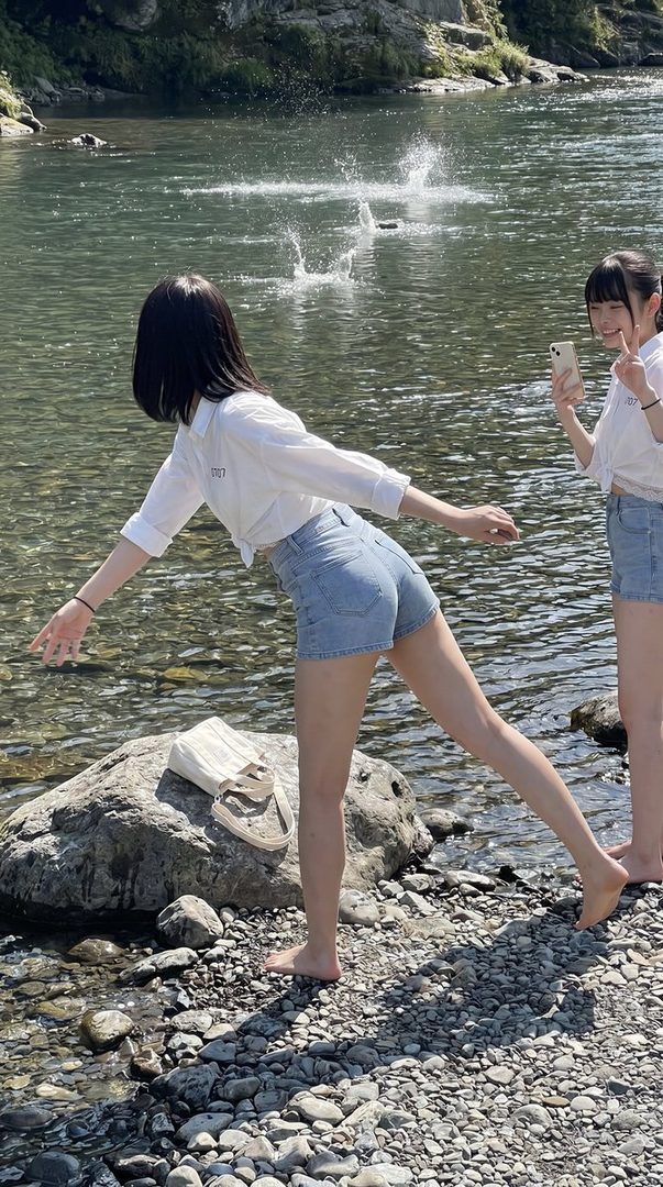 Two young women in casual summer outfits by a river, one throwing stones into the water, the other taking a photo with her phone.