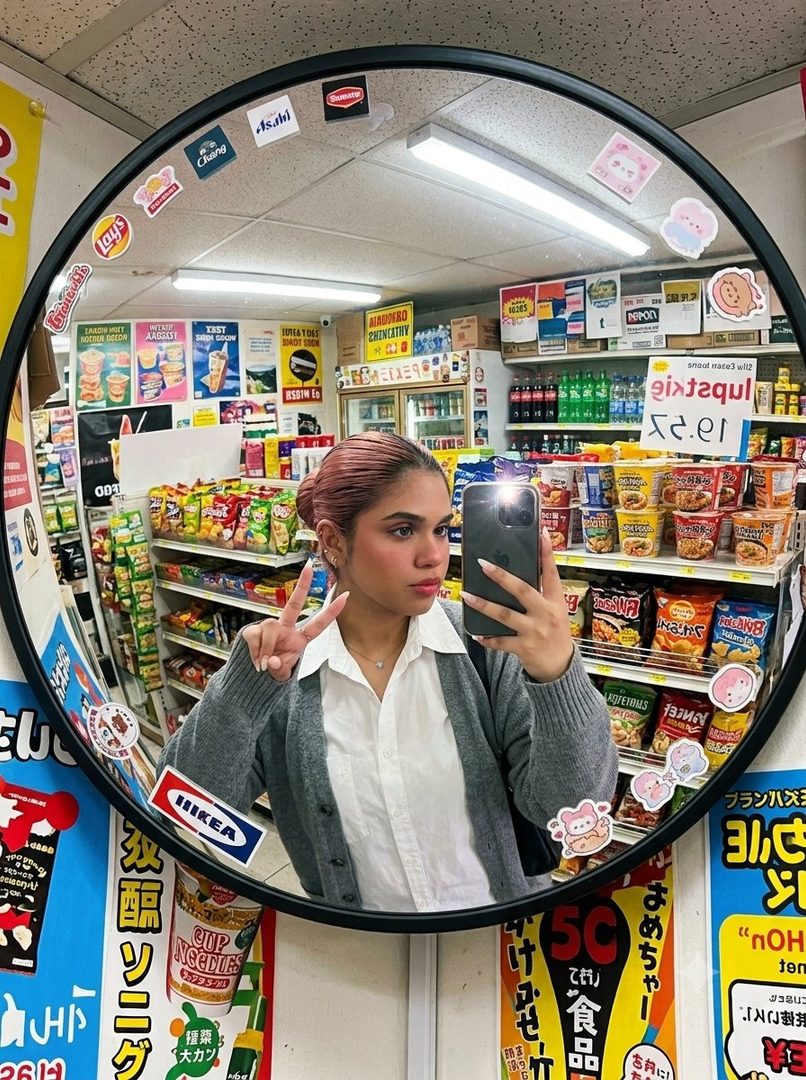 A young woman with pinkish-brown hair tied up takes a mirror selfie in a convenience store, wearing a white shirt and grey cardigan, and making a peace sign.