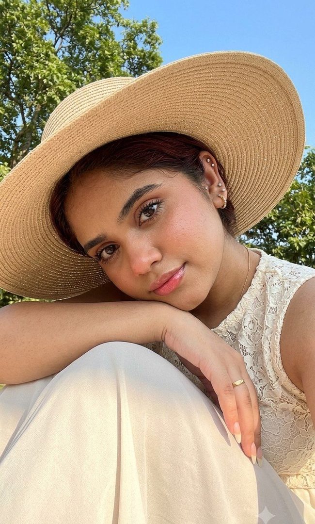A young woman with dark hair, wearing a wide-brimmed straw hat and a lace top, poses for a close-up selfie outdoors on a sunny day.