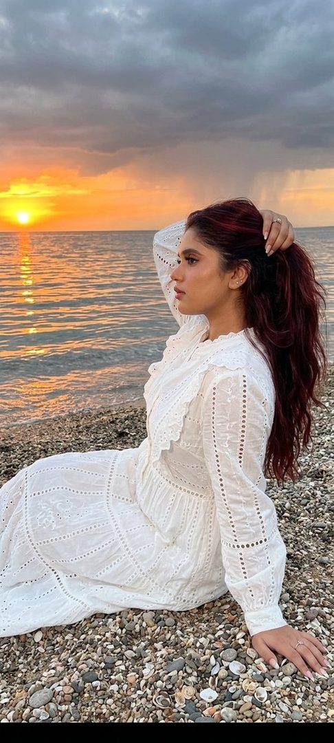 A woman with long red hair in a white dress sits on a pebble beach at sunset, looking out at the ocean.