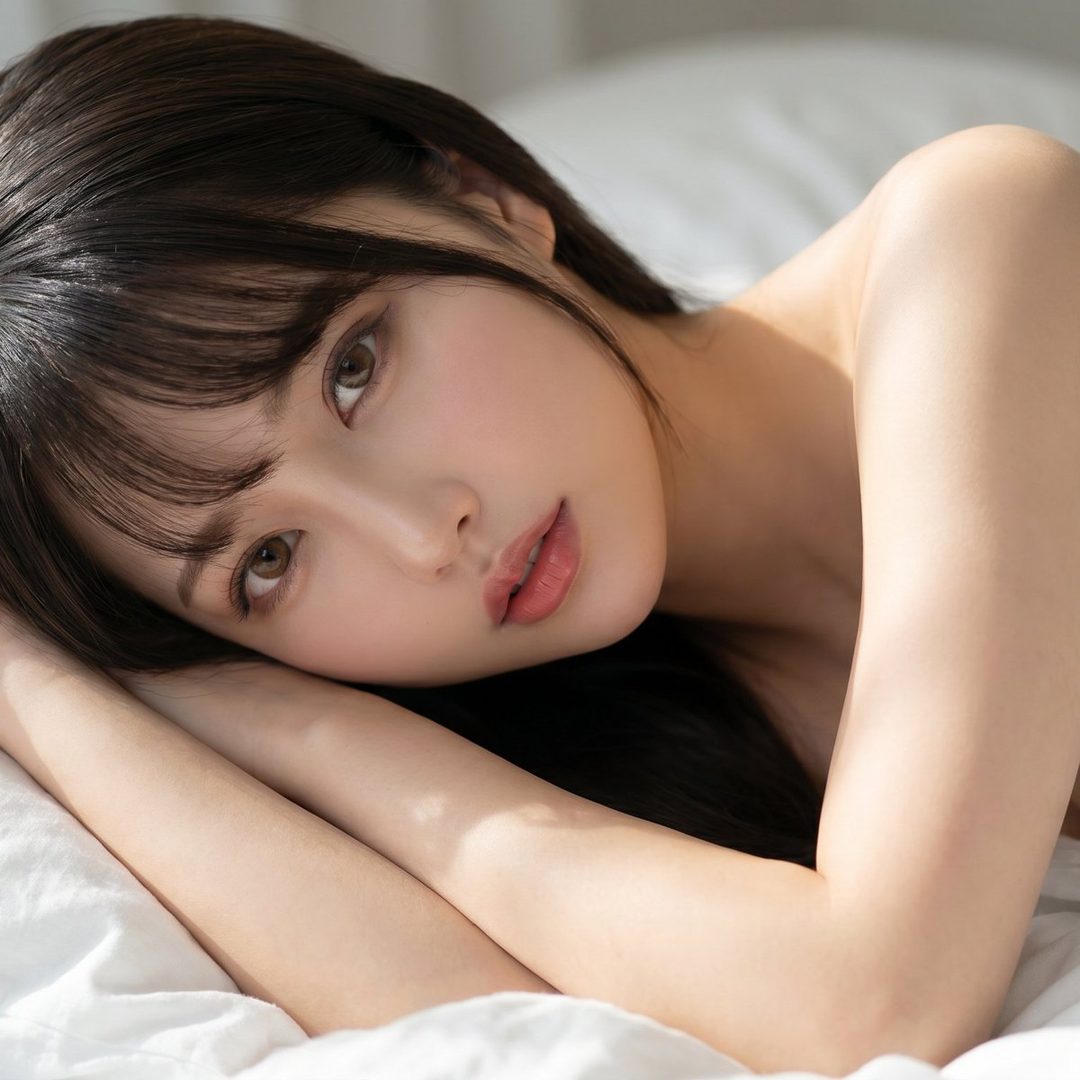A beautiful woman with dark hair and bangs lies on a white bed, looking suggestively at the camera with soft natural light on her skin.