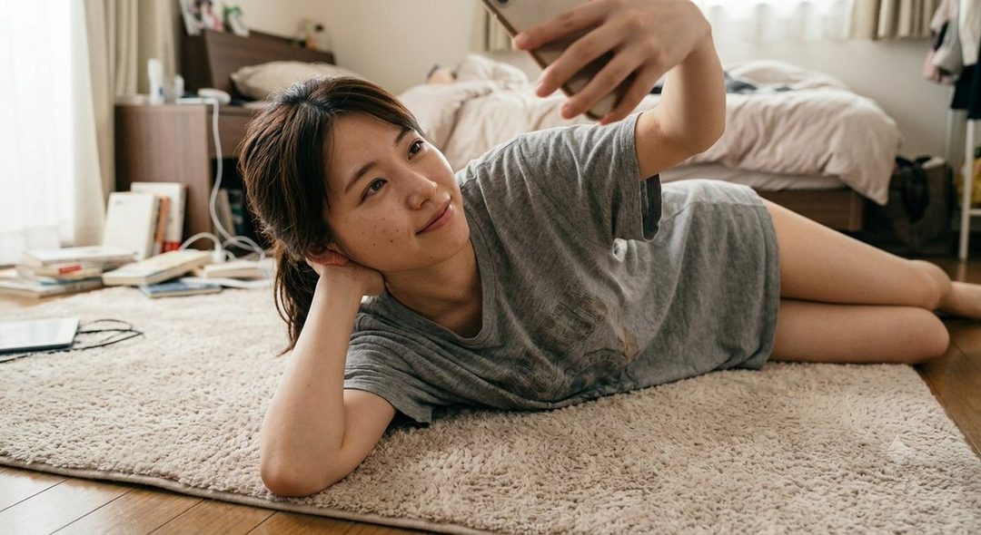 A young Japanese woman with freckles lies on a fluffy rug in a bedroom, taking a selfie with her phone.