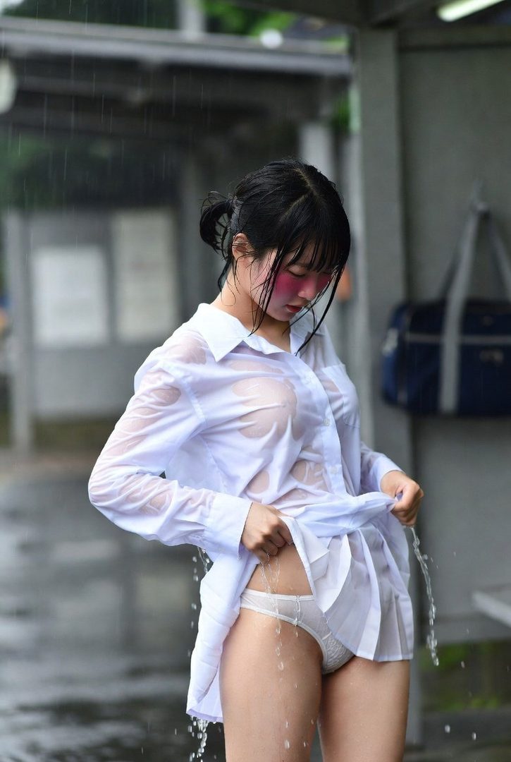 A young woman in a wet white shirt and white underwear stands in the rain, lifting her shirt.