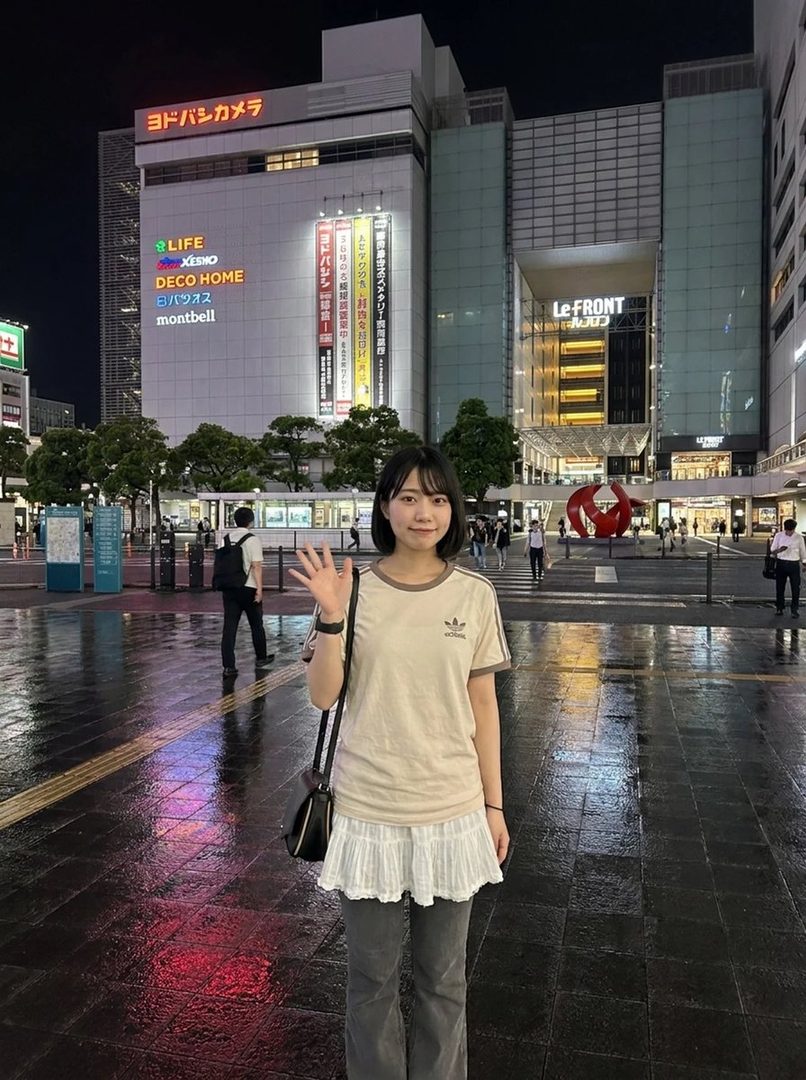 A young woman with short black hair waves at the camera in a city street at night, with a large department store in the background.