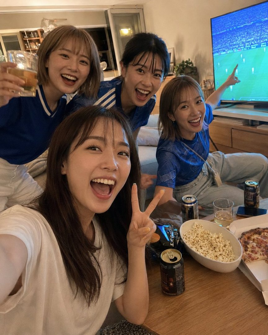 Four young women are excitedly watching a soccer game on TV in a living room, with snacks and drinks on the table.
