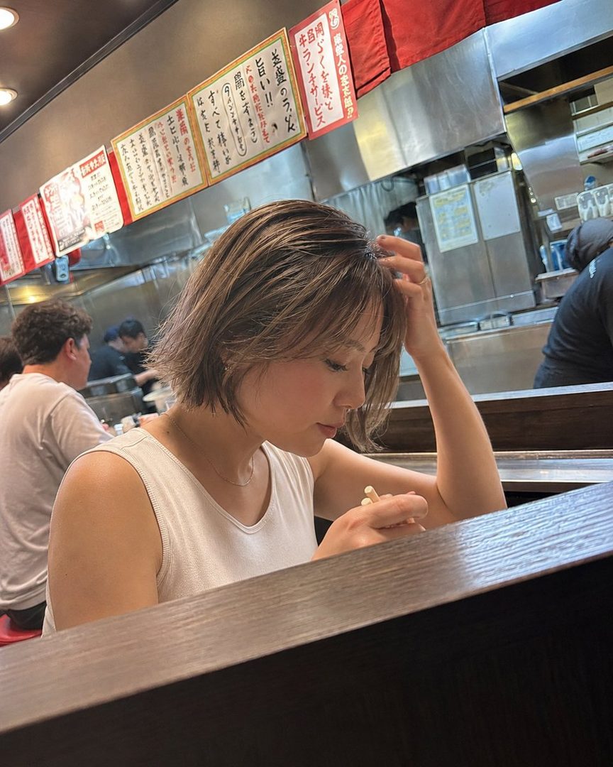 A woman with short brown hair is seen from the side, looking down while eating at a ramen shop counter, with Japanese signs in the background.