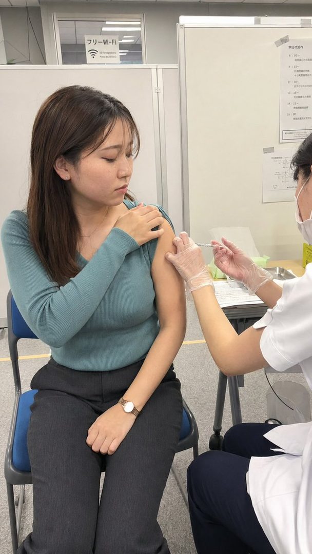 A young East Asian woman receives a vaccine injection in her arm while sitting in a chair indoors.