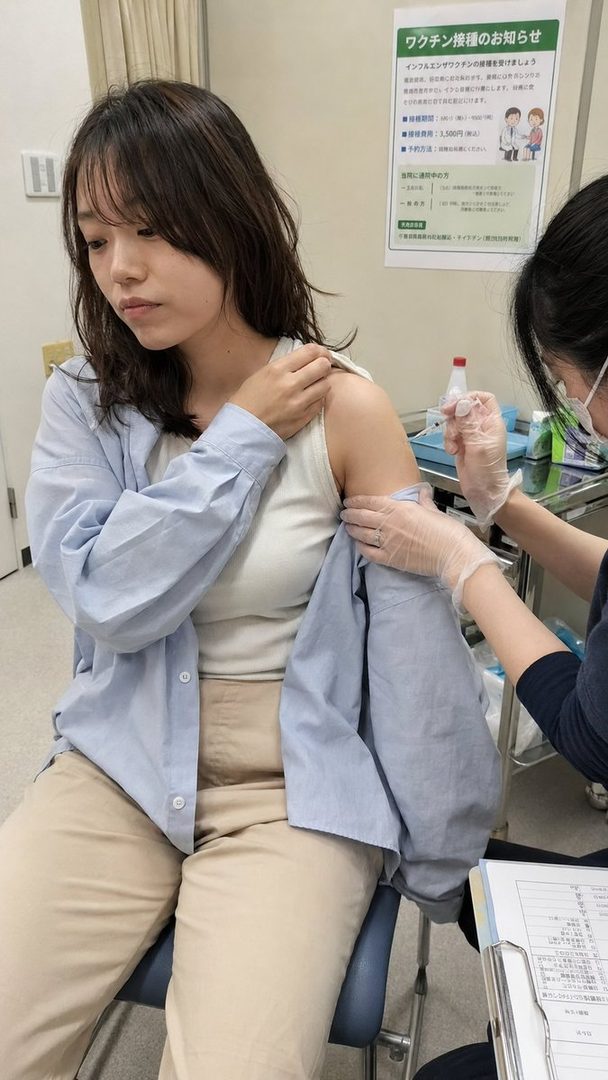 A young woman receives a vaccine injection in her arm while sitting in a medical setting. She is wearing a light blue shirt over a white tank top and beige pants. A medical professional in gloves is administering the shot.