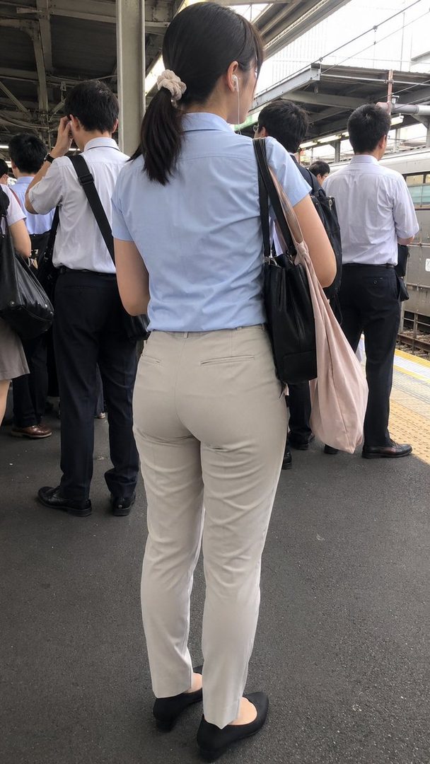 A woman in a light blue shirt and beige pants stands on a train platform, holding a bag.