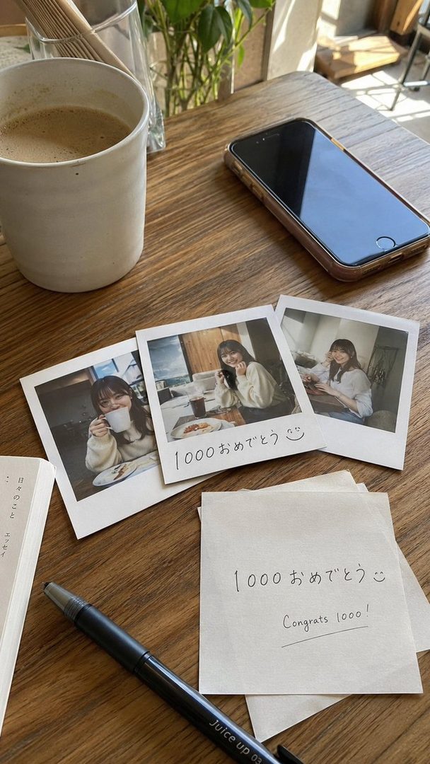 Three instant photos of a young East Asian woman smiling and holding a coffee cup, placed on a wooden table next to a coffee cup, a smartphone, a book, and a pen. A handwritten note says 'Congrats 1000!'.