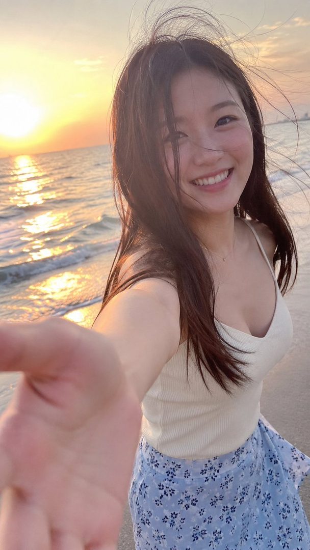 A young East Asian woman smiles at the camera on a beach at sunset, holding the camera out to take a selfie.