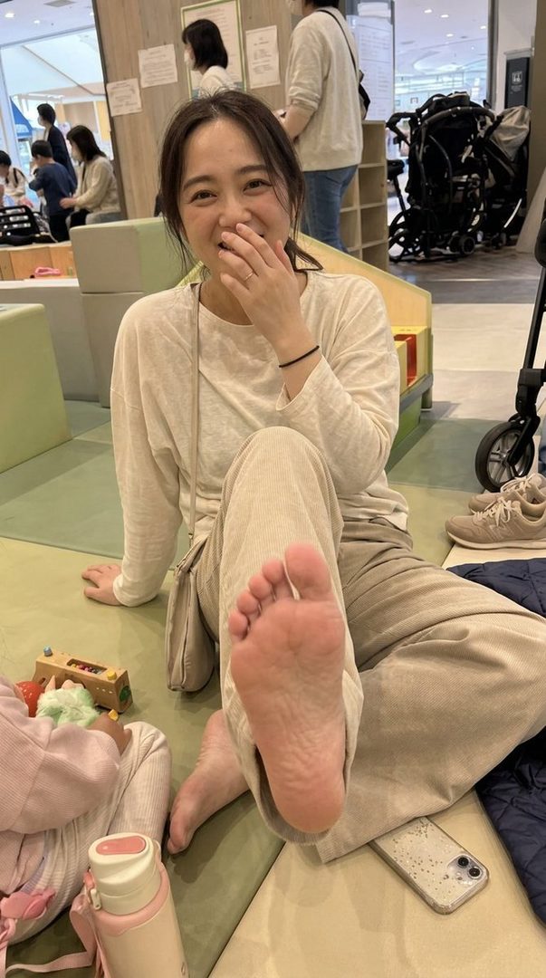 A smiling woman sits on the floor, showing her bare foot, with a baby and toys nearby.