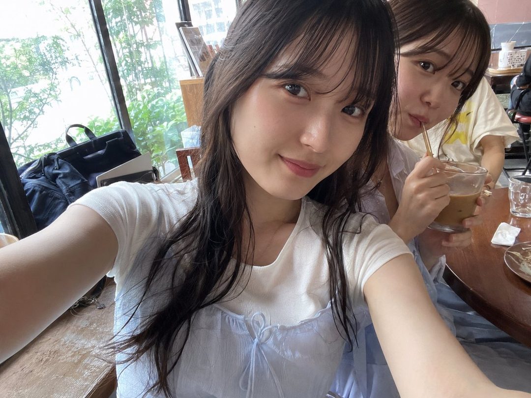 Two young women are taking a selfie in a cafe, one smiling at the camera while the other sips a drink.