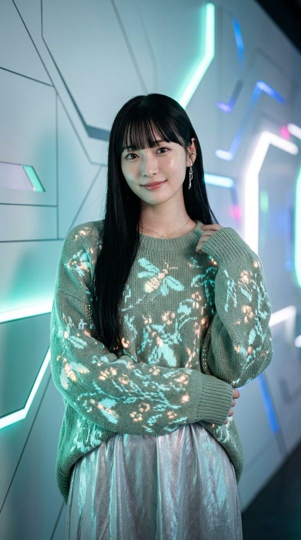 A young East Asian woman with long black hair and bangs, wearing a green sweater with a bee and floral pattern and a silver skirt, posing indoors with a futuristic background.
