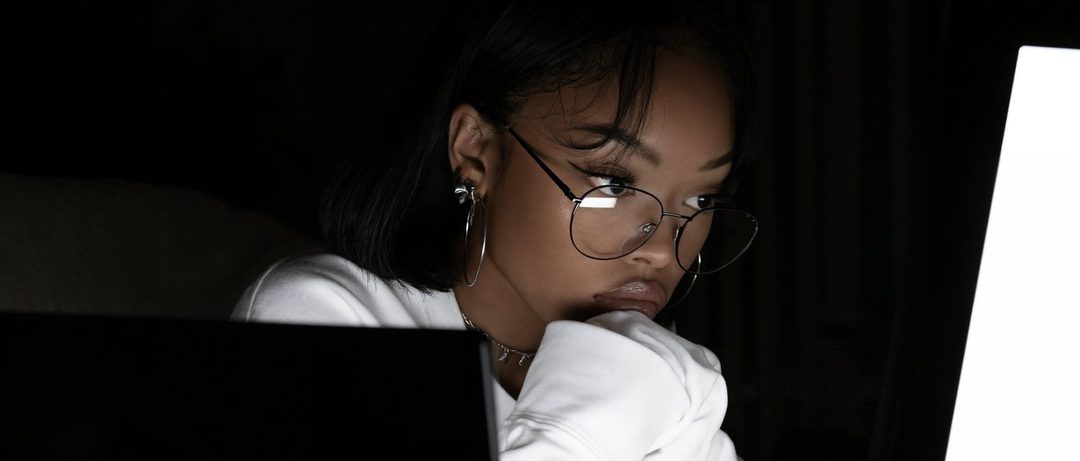 A young woman with glasses and dark hair is intently looking at a bright screen in a dark room.
