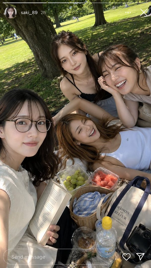 Four young women are enjoying a sunny picnic in a park, with one wearing glasses and holding a book, surrounded by fruits and drinks.