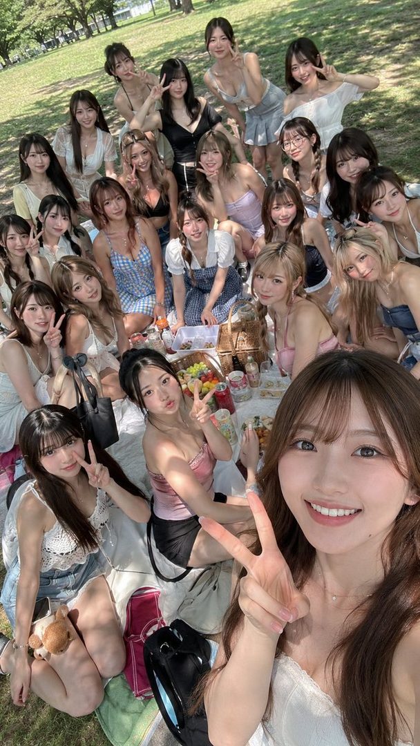 A large group of young women are enjoying a sunny outdoor picnic in a park, with many smiling and making peace signs for a group selfie.