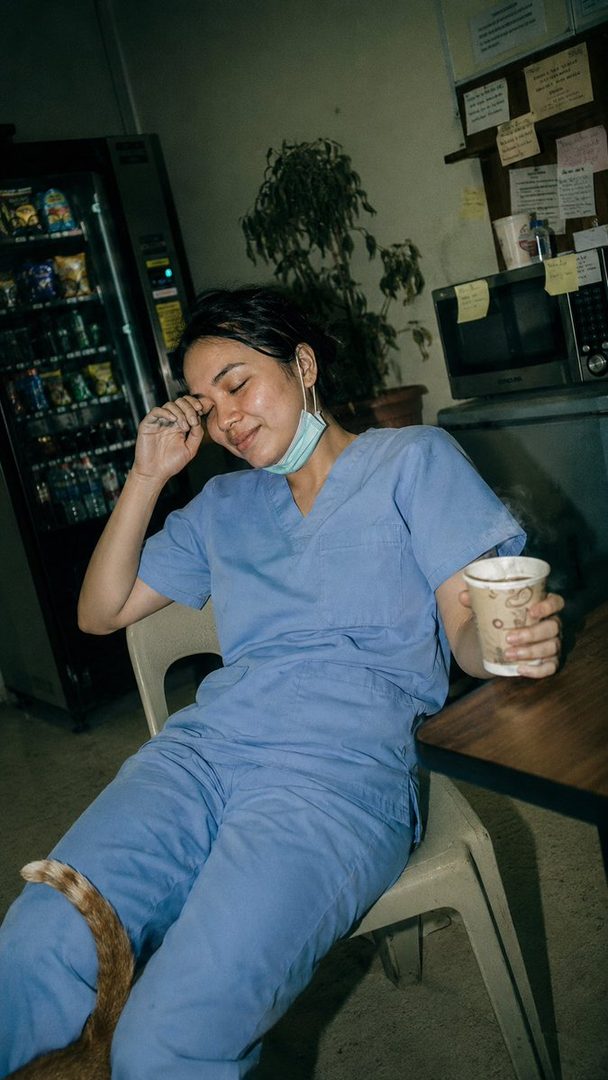A tired Indonesian nurse in blue scrubs sits in a hospital break room, holding coffee and rubbing her eyes, with a cat tail visible.