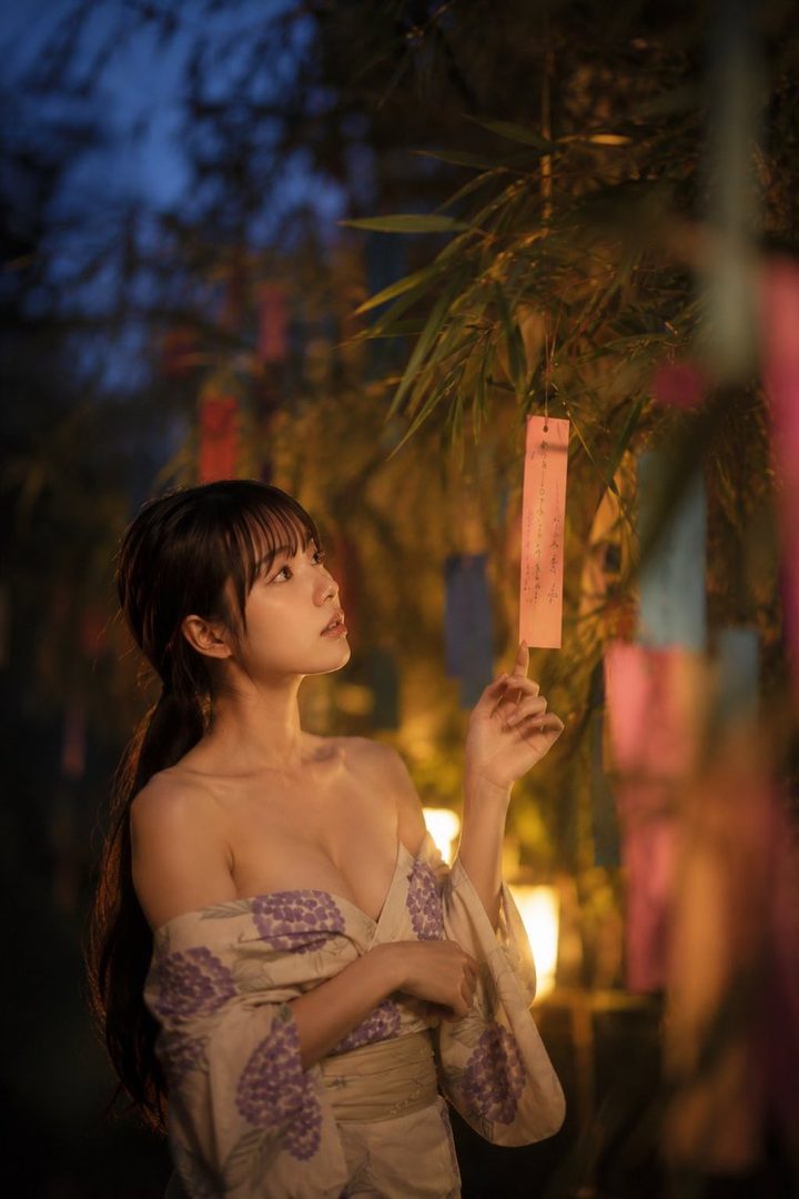 A young woman in a yukata looks up at a bamboo tree adorned with colorful tanzaku during a festival at night.
