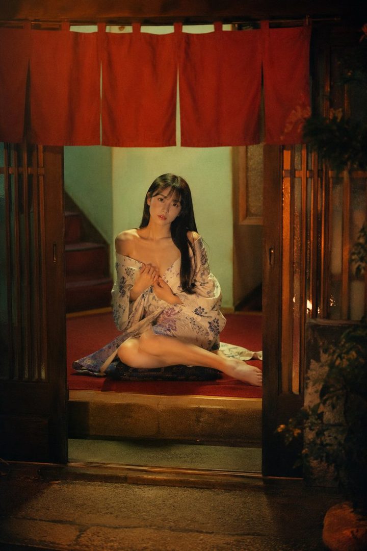 A young woman with long black hair sits indoors, wearing a floral robe that is partially open, revealing her chest. The lighting is dim and atmospheric.