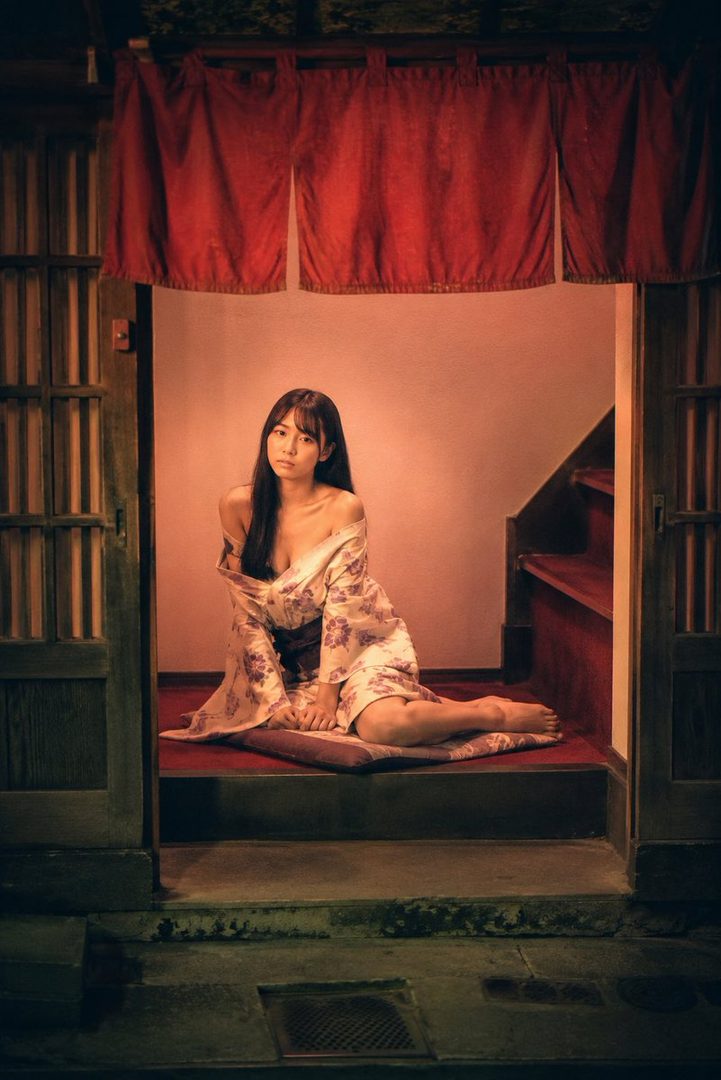 A young woman in a floral kimono sits on steps indoors, looking at the camera.