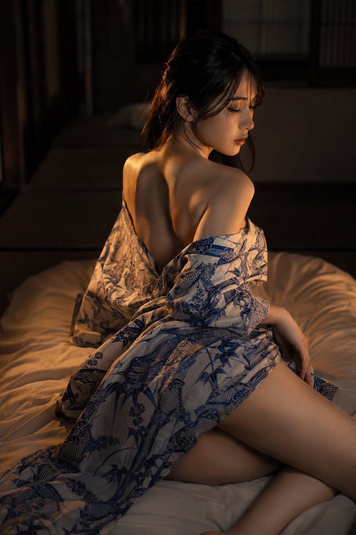 A young woman with dark hair, wearing a blue and white patterned robe, poses sensually on a bed in a dimly lit room.