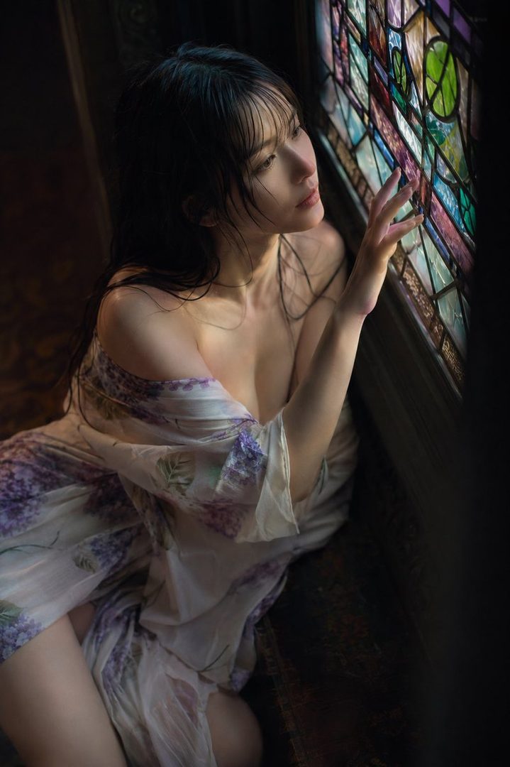 A young woman with long black hair gazes out a stained-glass window, wearing a floral robe.