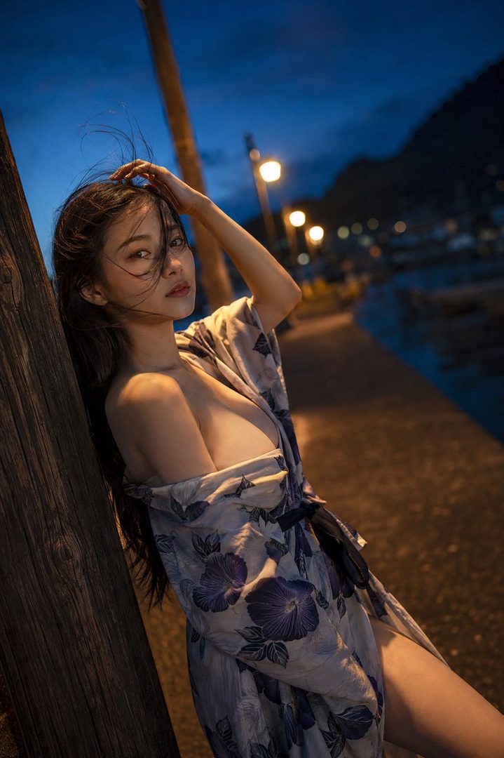 A beautiful East Asian woman in a floral yukata poses suggestively at night by a wooden post, with blurred city lights in the background.