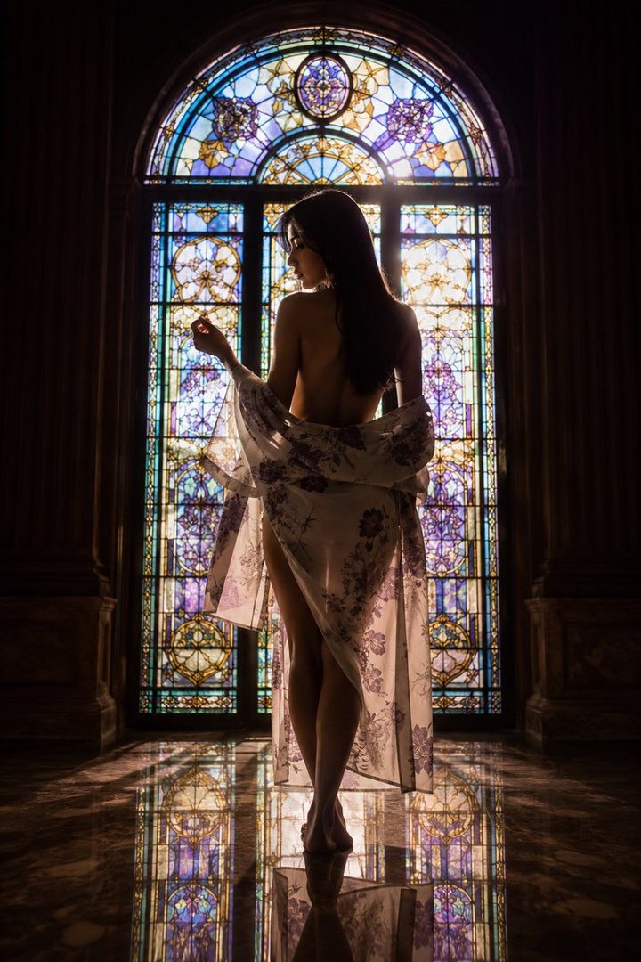 A woman in a floral robe stands in front of a stained glass window, illuminated by sunlight.