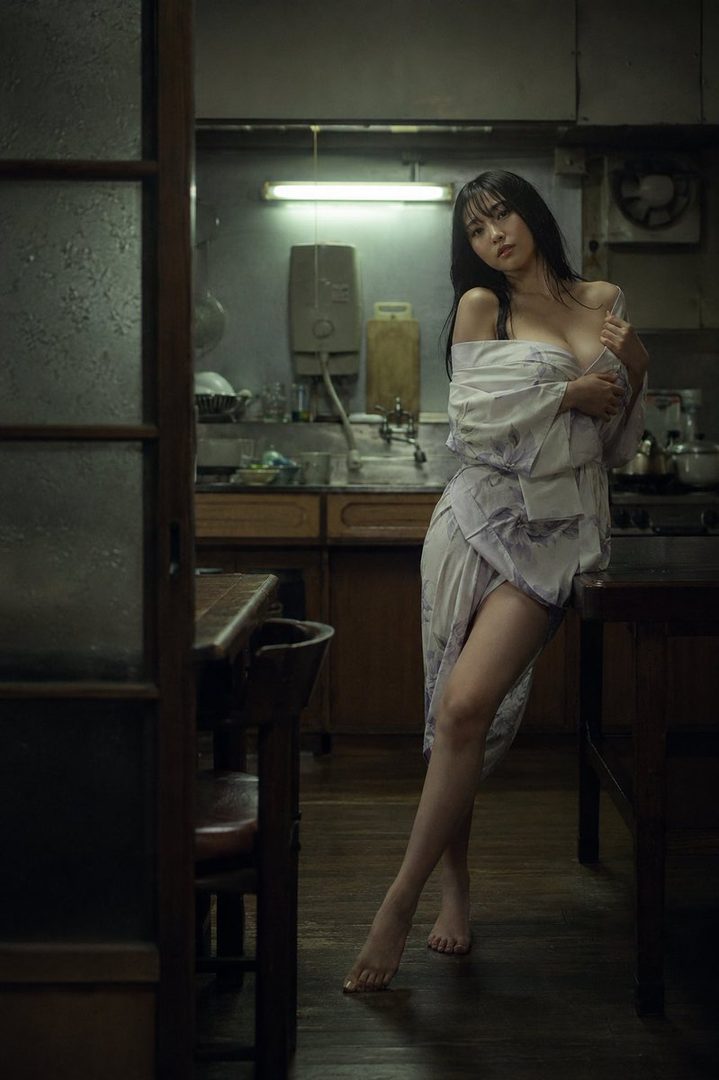 A young Japanese woman in a light-colored yukata poses suggestively in a dimly lit, vintage kitchen.