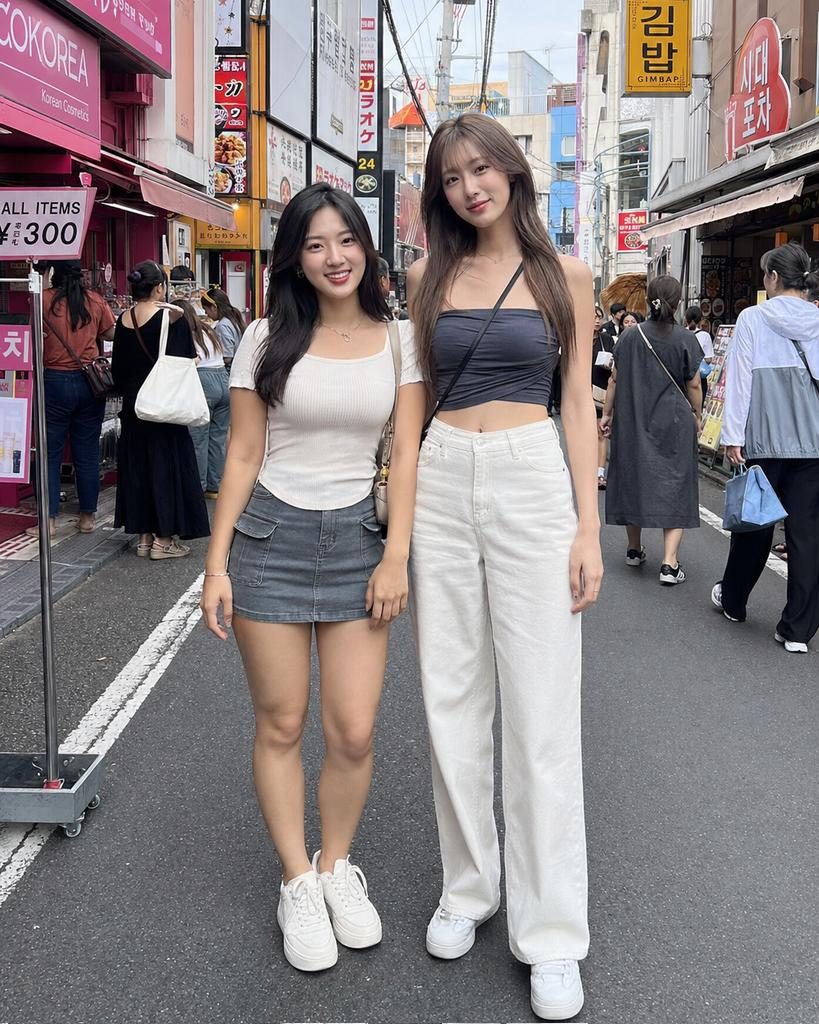 Two young women are standing on a bustling city street, smiling at the camera. One wears a white top and denim skirt, while the other wears a dark crop top and white wide-leg pants.