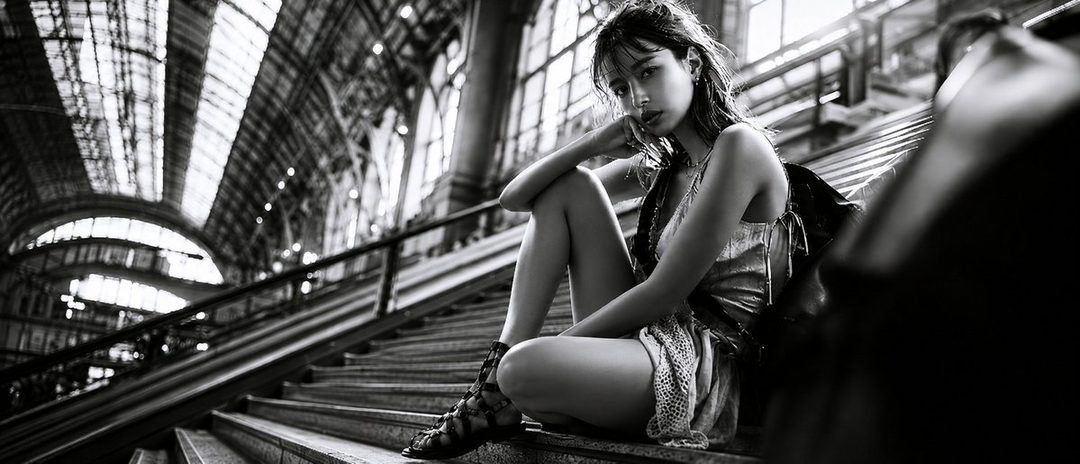 A young woman with long brown hair sits on steps in a grand, arched indoor space, looking directly at the camera in a black and white photograph.