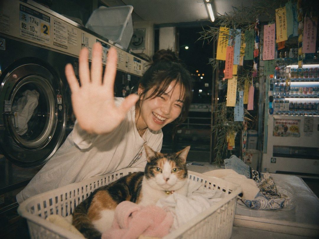A young woman with her hair in a bun smiles and waves at the camera while sitting next to a cat in a laundry basket. The background shows a laundromat with washing machines and a vending machine.