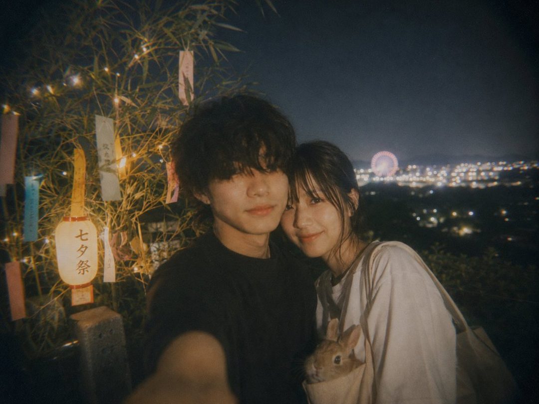 A young couple takes a selfie at night, with a decorated bamboo tree and a city view in the background.