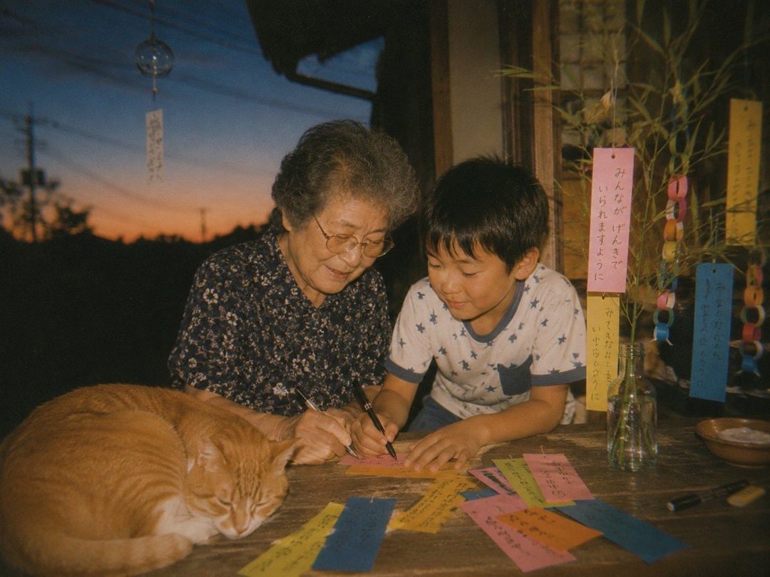 An elderly Japanese woman and a young boy write wishes on colorful strips of paper during the Tanabata festival, with a sleeping cat nearby.