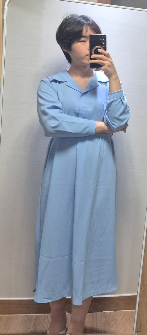A person with short black hair takes a mirror selfie, wearing a light blue long-sleeved dress indoors.