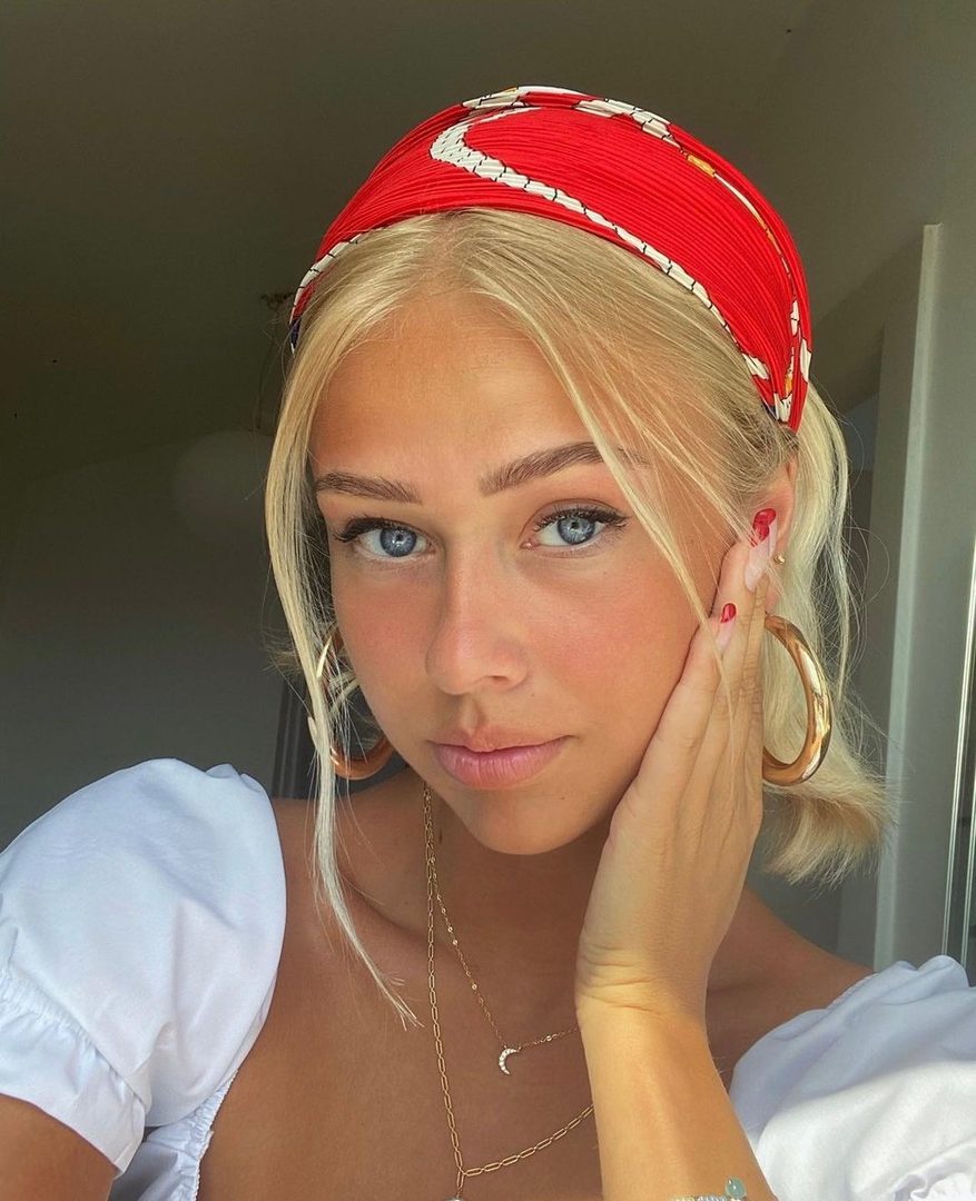 A young woman with blonde hair and blue eyes, wearing a red headband and a white top, poses for a selfie indoors.