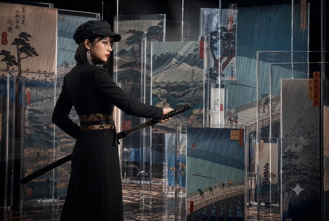 A woman in a black pinstripe outfit and a cap holds a katana, standing in front of a display of Japanese ukiyo-e prints depicting rainy scenes.